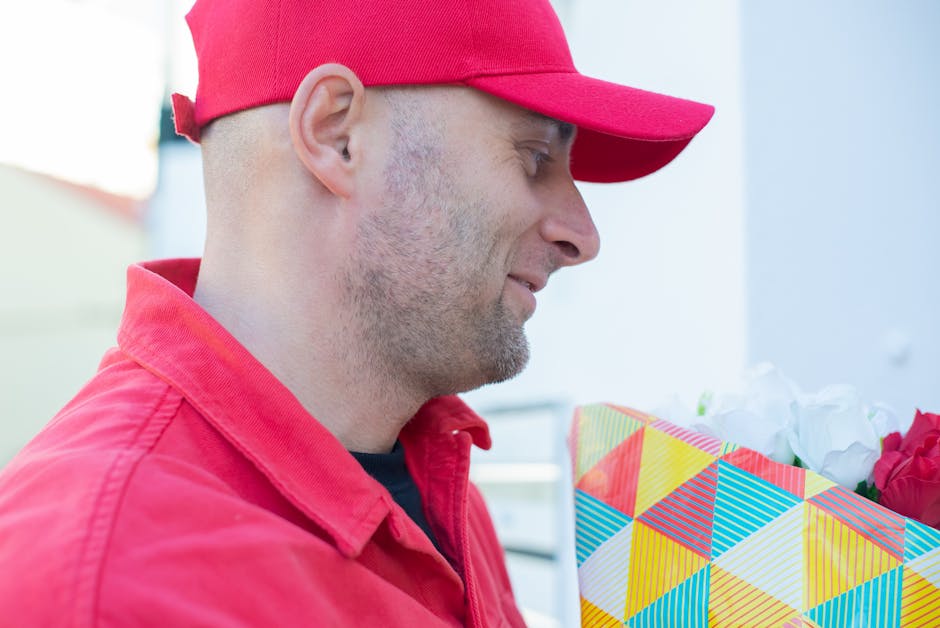 A man wearing a red baseball cap and a matching red jacket is holding a floral gift wrapped in colorful, geometrically patterned paper with vibrant yellow, blue, and red triangles. The bouquet contain