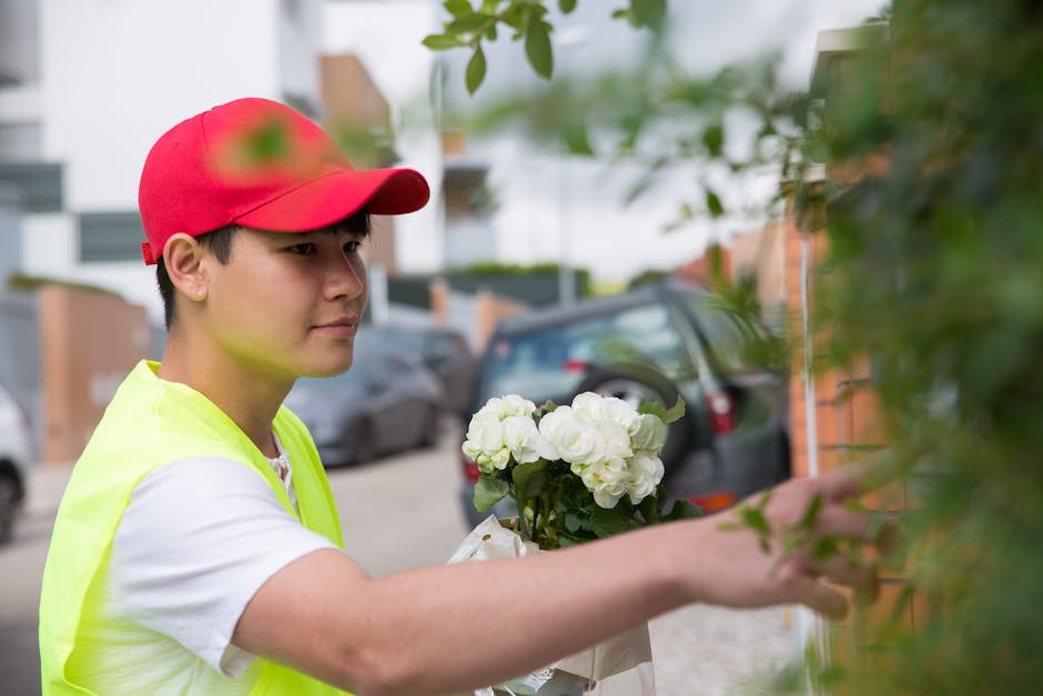 A young male flower delivery courier wearing a red cap and a bright yellow vest is taking a white floral bouquet, possibly consisting of roses or peonies, from a customer outside a residential area. T