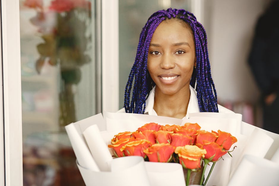 A smiling woman with long, purple braided hair holding a large bouquet of fresh orange roses wrapped in white paper. The bouquet is prominently displayed in front of her, with the petals showing vibra