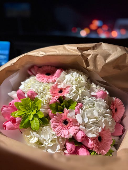 A bouquet of fresh flowers featuring white hydrangeas and pink gerbera daisies, accented with bright green foliage, wrapped in brown kraft paper with a partial view of a white tissue paper lining. The