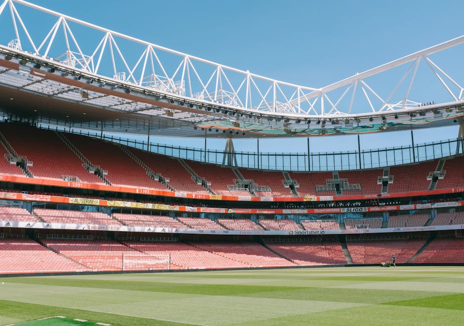 The image shows an empty football stadium with bright red seats arranged in multiple tiers around a lush, green grass pitch. The stadium features a modern white roof structure supported by large beams