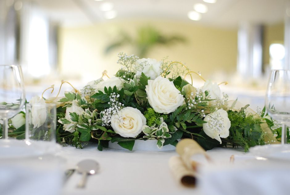 A floral arrangement featuring white roses, delicate baby's breath, and variegated green leaves, set in a white dish on a table. The bouquet is surrounded by clear wine glasses, a rolled napkin, and s