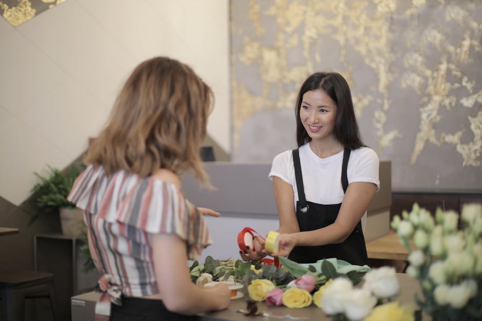 A florist at flowerstottenham.co.uk is engaging in a floral arrangement, holding a roll of red ribbon and a yellow tape dispenser while conversing with a customer. The florist, a young woman with long
