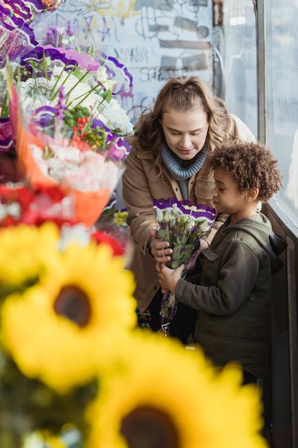 A woman and a young girl are inside a flower shop near a large window, surrounded by vibrant floral arrangements. The woman, with curly hair, is wearing a beige coat and is smiling gently as she holds