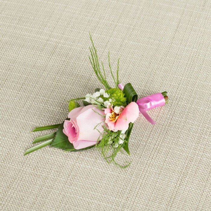 Carefully positioned on a softly textured light beige fabric-evoking the comfort of a Tottenham home-this refined wedding boutonniere captures the intricate beauty of locally made floristry. A single pink rosebud forms the elegant centerpiece, its petals curled in a spiral of gentle blush, edges fading to the palest rose, conjuring a sense of serene freshness as if chosen from a sunlit garden beside Downhills Park. Flanking the rose is a soft alstroemeria bloom in blush and peach tones, its open petals revealing a sunburst of creamy yellow and delicate line markings that draw the eye inward. Complementing these focal flowers, clusters of white baby's breath intersperse airy softness, their miniature blooms like clouds drifting above the greenery. Accents of petite, spherical green chrysanthemums add both lively contrast and a playful pop of moss-green texture, grounding the arrangement in nature. The boutonniere is enriched further by finely arching fern fronds and slender blades of dark foliage, echoing the elegant wildness seen in local gardens, while broader emerald leaves provide form and structure at the base-some leaves artistically folded for added dimension. The bundled stems are wrapped tightly in a glossy fuchsia satin ribbon tied into a crisp, celebratory bow, its sheen adding a burst of joy reminiscent of Tottenham's community festivals. Resting at a gentle diagonal, the arrangement is bathed in soft, natural light, illuminating each contour, colour, and texture. Perfect for a special event-a Tottenham wedding or milestone celebration-this boutonniere exudes romance, sophistication, and handcrafted local care.