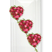 The photograph captures an elegant and emotionally charged triple-heart floral arrangement, thoughtfully designed as a funeral or memorial tribute. Three hearts, each exquisitely sculpted from a dense mass of roses, descend diagonally in a vertical line, creating a visual rhythm that naturally draws the eye from top to bottom. The roses cover the entire surface of each heart, with velvety crimson blooms interspersed with brighter pinks and gentle blush tones, forming a rich tapestry of colour associated with deep love, affection, and gratitude. Certain flowers are still in bud, tight and smooth, while others unfurl in layered whorls that add depth and softness, offering a sense of movement and life within the stillness of the arrangement. Tracing the outline of each heart, fine strands of green ivy curve and twist, their small, glossy leaves lending a fresh woodland touch that softens the boldness of the roses. The ivy trails from one heart to the next, visually binding them together and evoking themes of family connection, shared memories, and unbroken bonds. Set against a crisp white background, which might recall the clean walls of a modern funeral chapel or a serene space in Tottenham where loved ones gather, the colours appear exceptionally vivid and pure. The lighting is gentle yet direct enough to highlight the natural sheen on the leaves and the plush texture of each petal. This arrangement feels particularly suited to a dignified farewell in Tottenham, offering a graceful, symbolic expression of enduring love that can accompany a casket, rest beside a photograph, or stand near the altar during a service of remembrance.